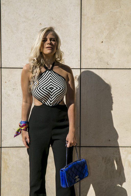 Mulher encostada em uma parede de pedra, vestindo um top frente única preto e branco e calças pretas, segurando uma bolsa de mão azul.