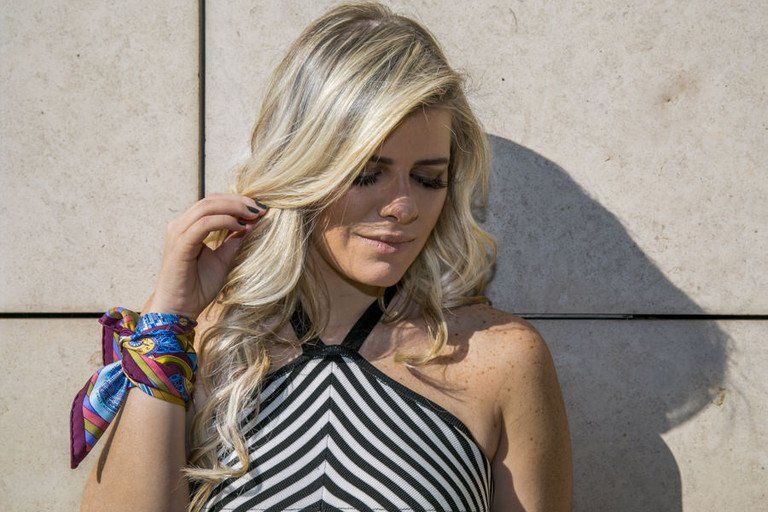 Mulher loira com blusa tomara que caia preta e branca, em pé junto a um muro de pedra, prendendo o cabelo com pulseiras coloridas.