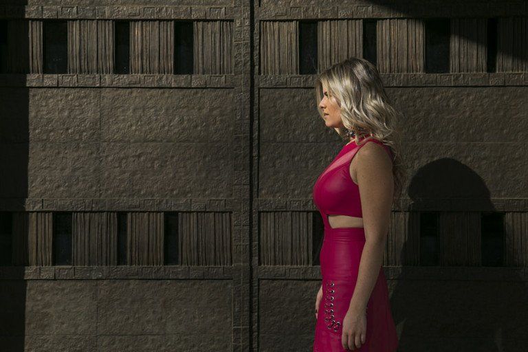 Mulher de vestido magenta em pé, de perfil, junto a uma parede de concreto texturizado.