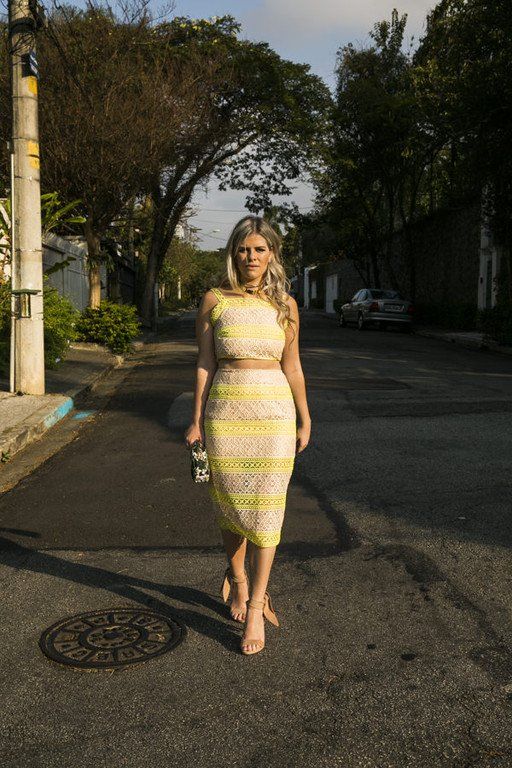Mulher com um vestido amarelo listrado em pé numa rua residencial ensolarada.