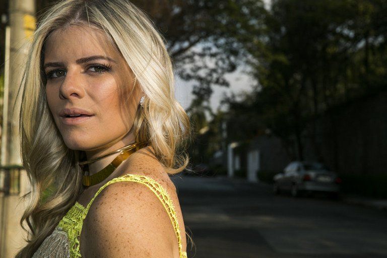 Mulher loira de vestido amarelo em pé ao ar livre numa rua arborizada, olhando para a câmera.