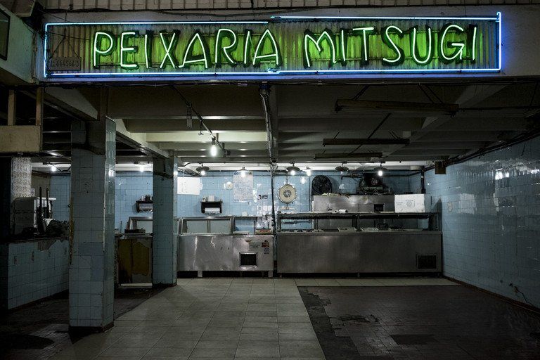 Fachada da loja Paxaria Mitsugi iluminada com luzes de néon, interior vazio e equipamentos de cozinha em aço inoxidável.