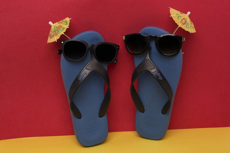 Chinelo azul com óculos de sol pretos e detalhes florais amarelos sobre um fundo vermelho e amarelo.
