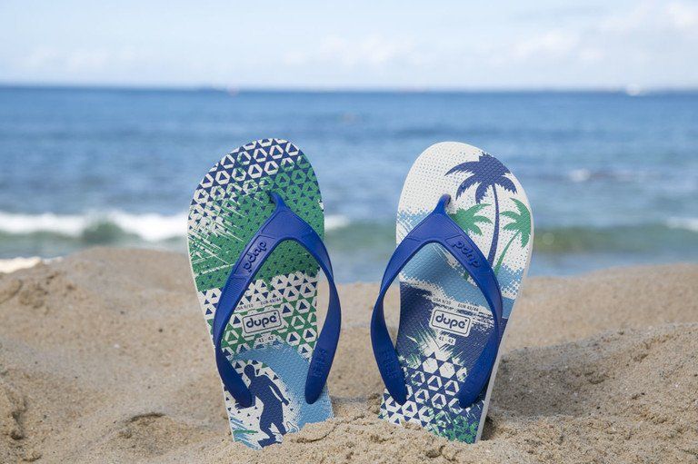 Duas sandálias de dedo estampadas na areia à beira-mar, com ondas e céu azul ao fundo.
