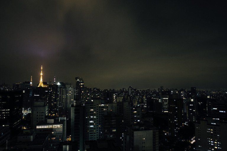 Horizonte escuro da cidade à noite, com arranha-céus iluminados e uma torre brilhante sob um céu nublado.