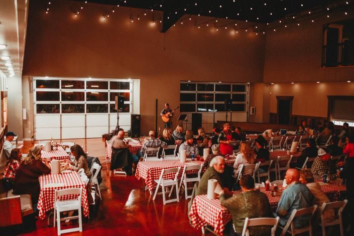 Indoor event hall with guests seated at red-and-white checkered tables under string lights