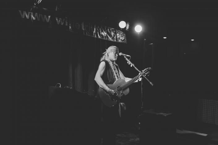 Black-and-white photo of a musician singing and playing guitar onstage under bright lights