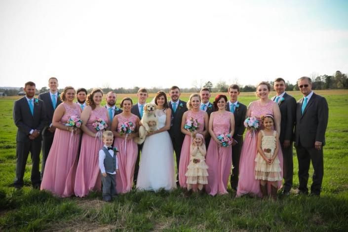Wedding party posing outdoors on a grassy field, with bridesmaids in pink dresses and a bride in white.