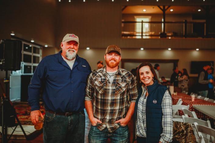 Three people smiling indoors at a rustic event venue with warm lights and wooden railings.