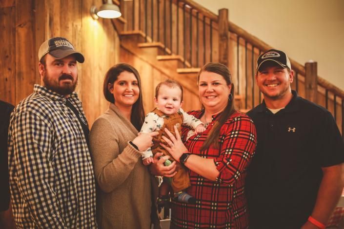 Five people smiling together indoors in front of a wooden staircase, holding a baby.