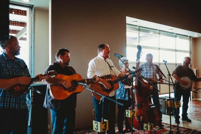 Bluegrass band performing indoors with guitars and a cello beside large windows