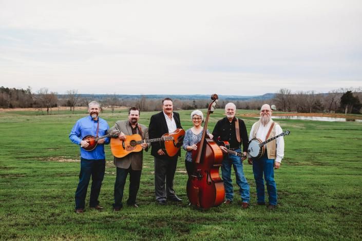 Five musicians stand outdoors in a grassy field holding guitars, fiddle, and upright bass under a cloudy sky.