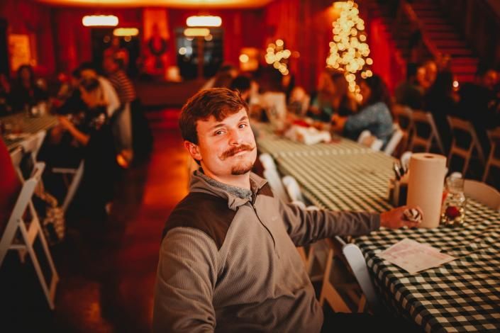 Man sitting at a checkered table in a warmly lit restaurant, smiling at the camera with a mug nearby