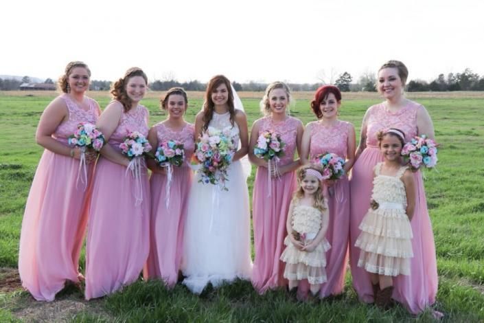 Bridal party in pink dresses and a white gown posing in a grassy field with bouquets