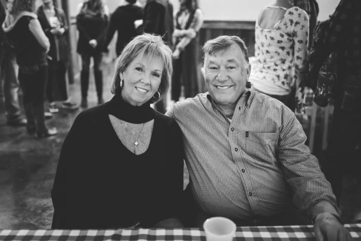 Two smiling people seated at a table in a crowded indoor event, with a checkered tablecloth and cup in front.