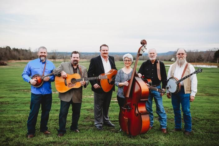 Six musicians with instruments posing in a grassy field under a cloudy sky
