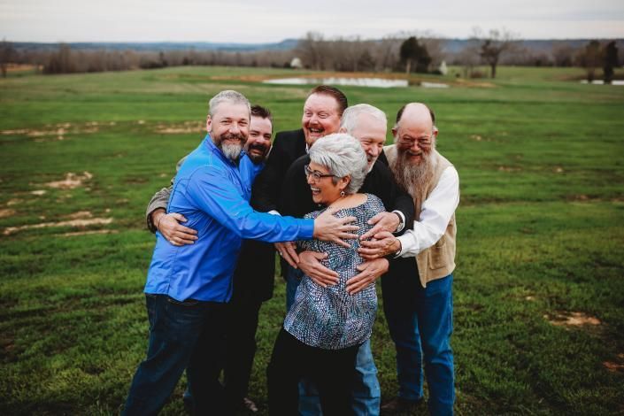 Six people hugging in a grassy field under an overcast sky