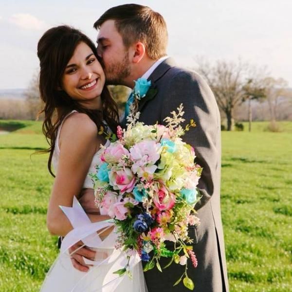Wedding couple in a grassy field, groom kissing bride on the cheek as she holds a colorful bouquet.