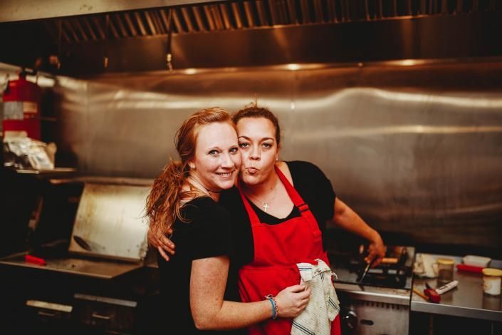 Two smiling restaurant workers in a stainless-steel kitchen, one in a red apron.