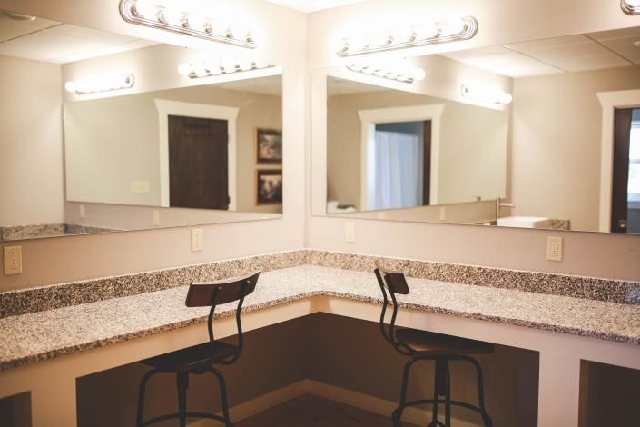 Mirrored vanity with lit bulbs, granite countertops, and two black stools in a dressing room