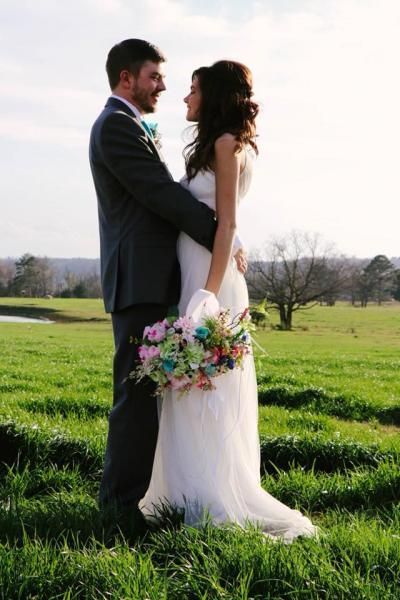 Bride and groom embracing outdoors in a grassy field, bride holding a colorful bouquet