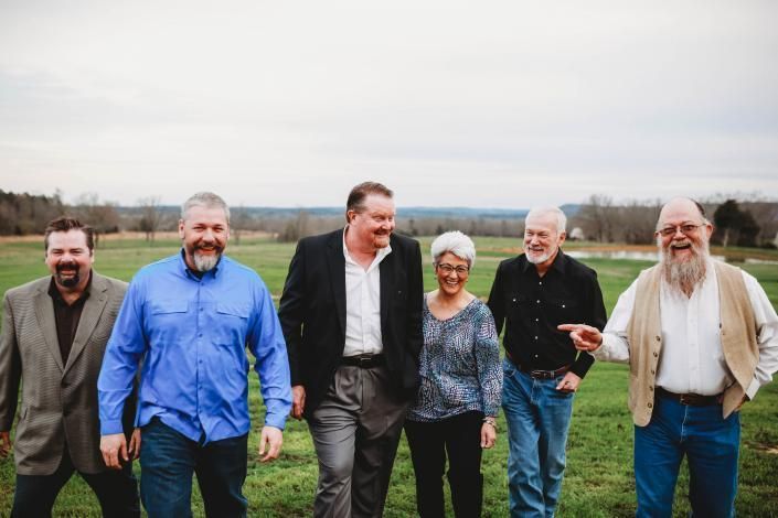 Six people walking together on a grassy field, smiling under an overcast sky.