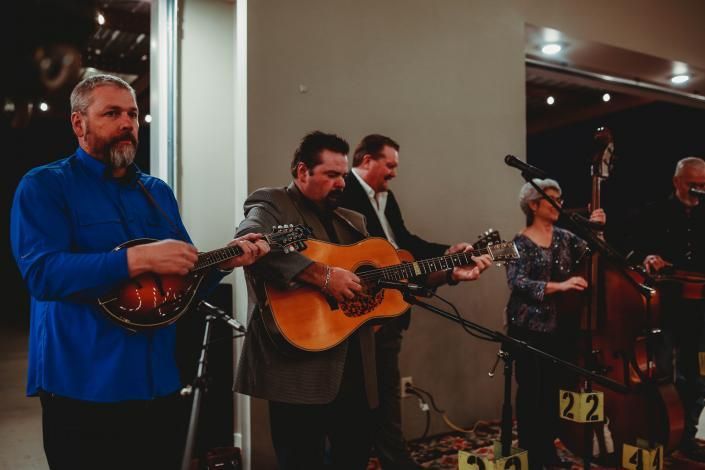 Four musicians performing indoors, with mandolin, guitars, and microphones on a stage.