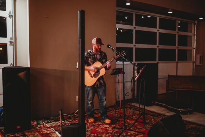 Musician playing acoustic guitar on a small indoor stage with microphones and speakers, in warm lighting