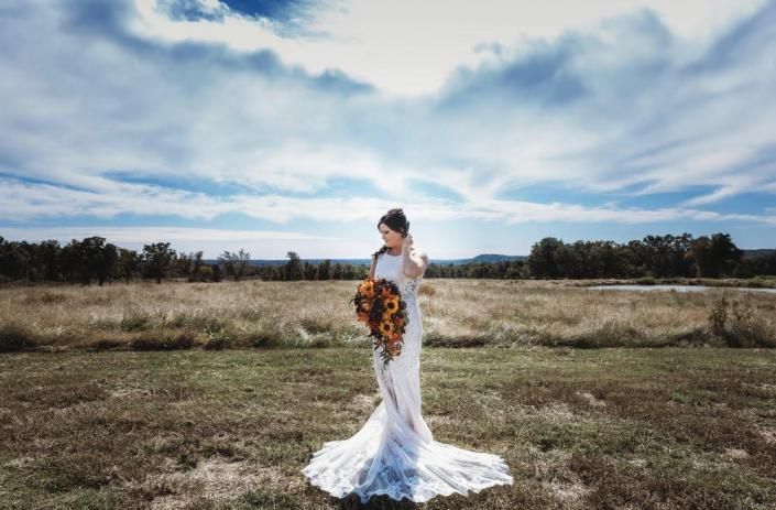 Bride in a white gown holding an autumn bouquet in a grassy field under a cloudy sky