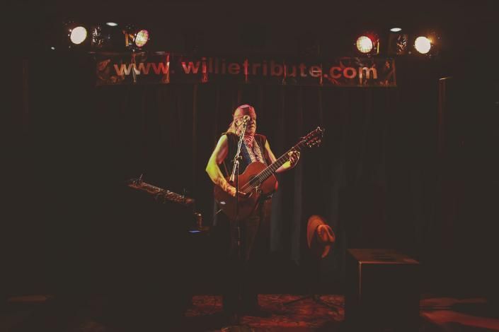 Solo musician on a dimly lit stage, playing guitar under warm spotlights, with a website banner overhead
