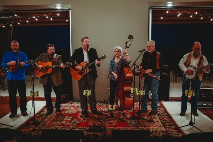 Six musicians perform on a small indoor stage with microphones and string instruments on patterned rugs.