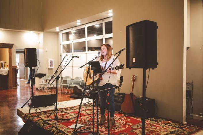 Musician performing with guitar and microphone on a rug in a living room, flanked by speakers and guitars.