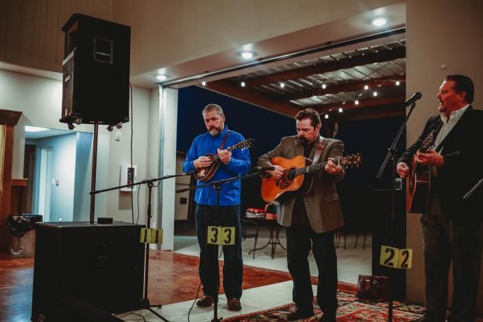 Three musicians perform on a small indoor stage with guitars and microphones under warm lights.