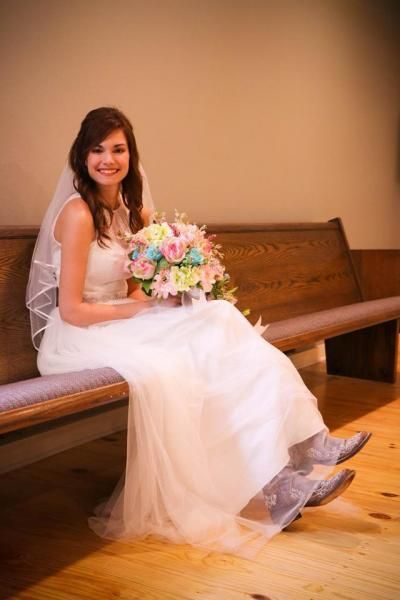 Bride in a white wedding dress sitting on a wooden bench, holding a pastel bouquet and smiling.