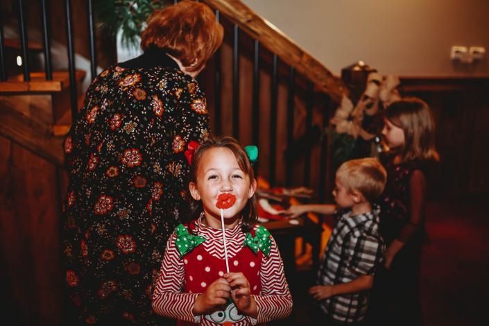 Child in a red holiday sweater smiles at the camera while holding a treat at a festive indoor gathering