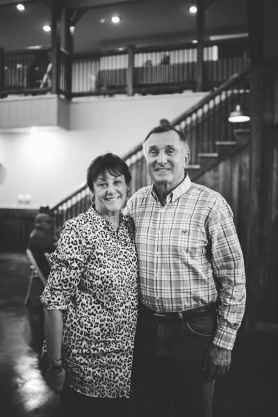 Two people smiling together indoors on a staircase landing, with railing and lights in the background.