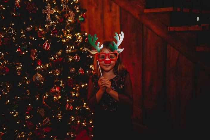 Child in reindeer antlers and red glasses smiling beside a lit Christmas tree