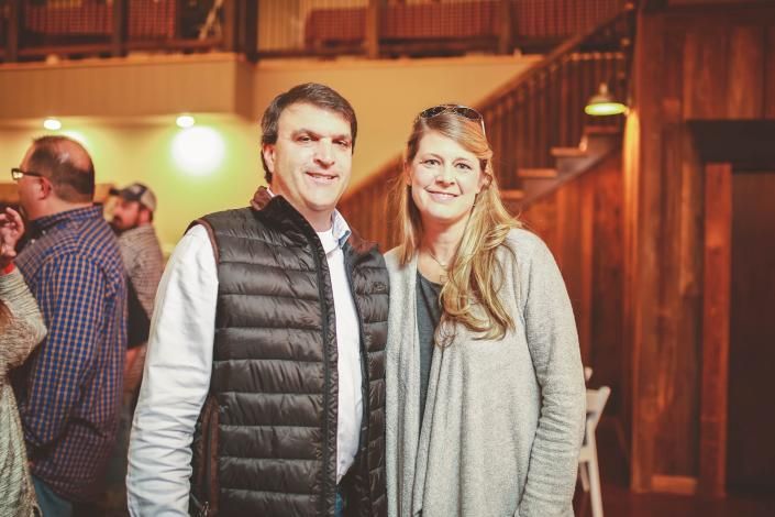 Two people smiling indoors at a warmly lit event, with wooden balconies in the background.