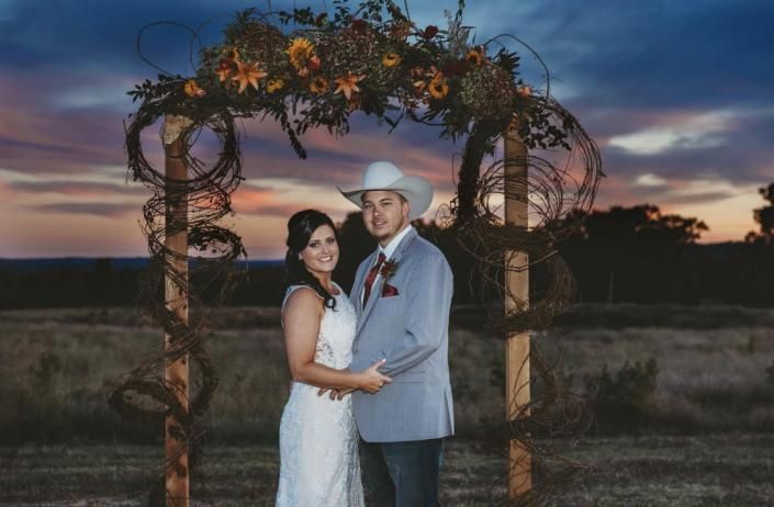 Couple in formal attire under a flowered arch at sunset in a field, holding hands and smiling
