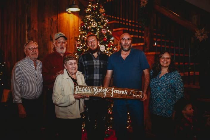 Six people posing in front of a lit Christmas tree, holding a wooden sign in a dim restaurant.