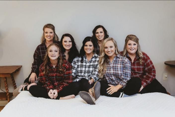 Seven people in plaid shirts sitting on a bed, smiling indoors.