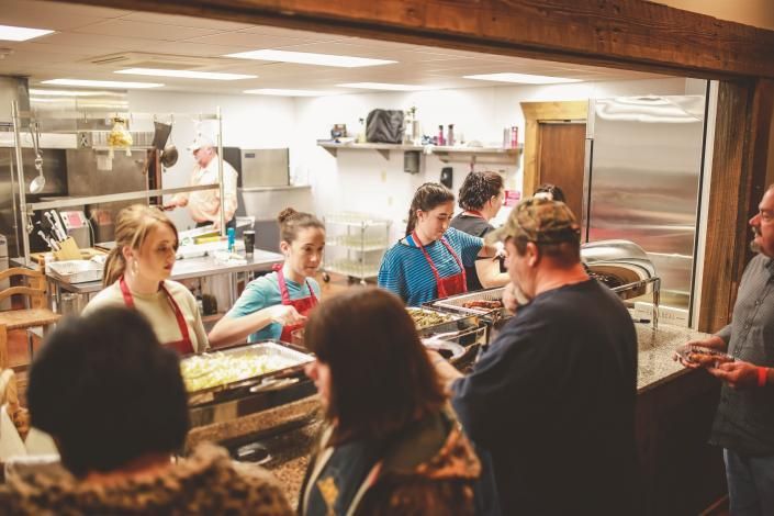 People serving food at a busy indoor market counter, with customers waiting and chatting.