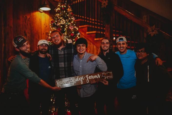 Six people holding a wooden sign in front of a Christmas tree on a warmly lit staircase