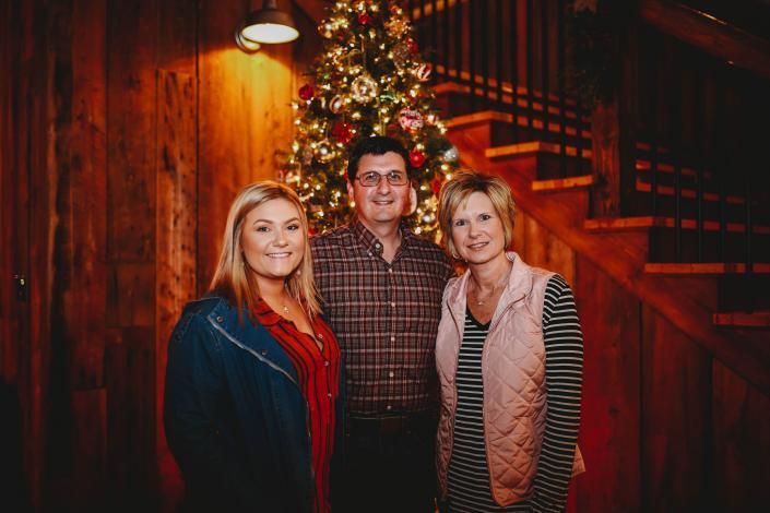 Three people standing by a decorated Christmas tree in a warmly lit wooden interior
