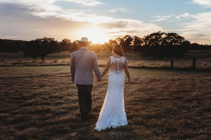 Couple walking hand in hand across a field at sunset, with trees silhouetted in the background.