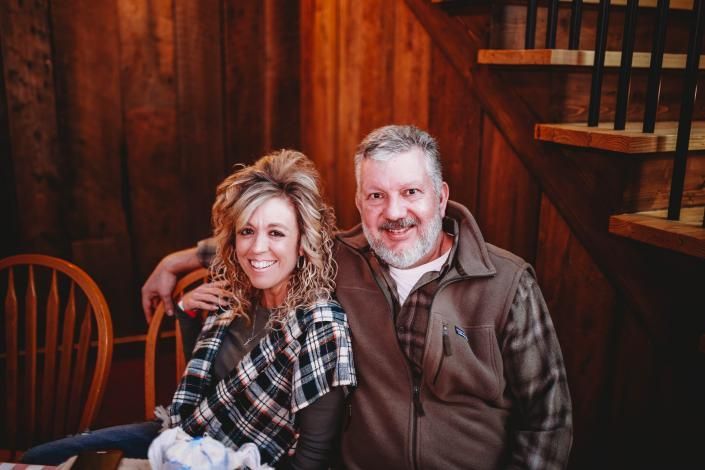 Smiling couple seated together in a rustic wooden interior, wearing plaid and brown jackets.