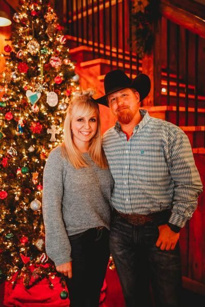 Couple posing beside a decorated Christmas tree and staircase, smiling in a warmly lit holiday setting