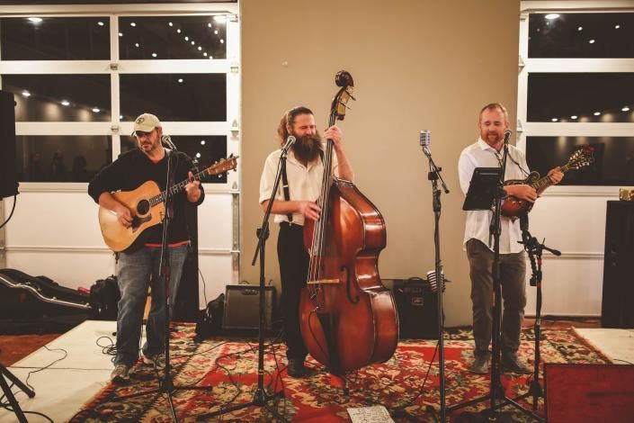 Three musicians perform in a warmly lit room, playing guitar, upright bass, and violin on patterned rugs.