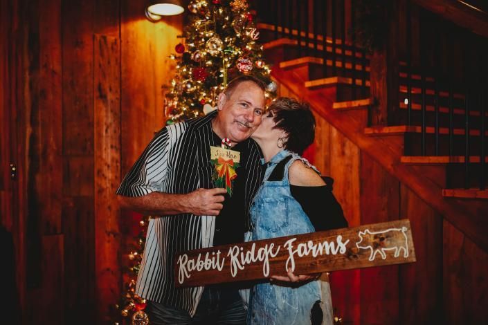 Couple posing by a Christmas tree on a wooden staircase, holding a holiday sign and flowers