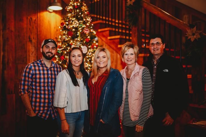 Five people posing indoors beside a lit Christmas tree and staircase.